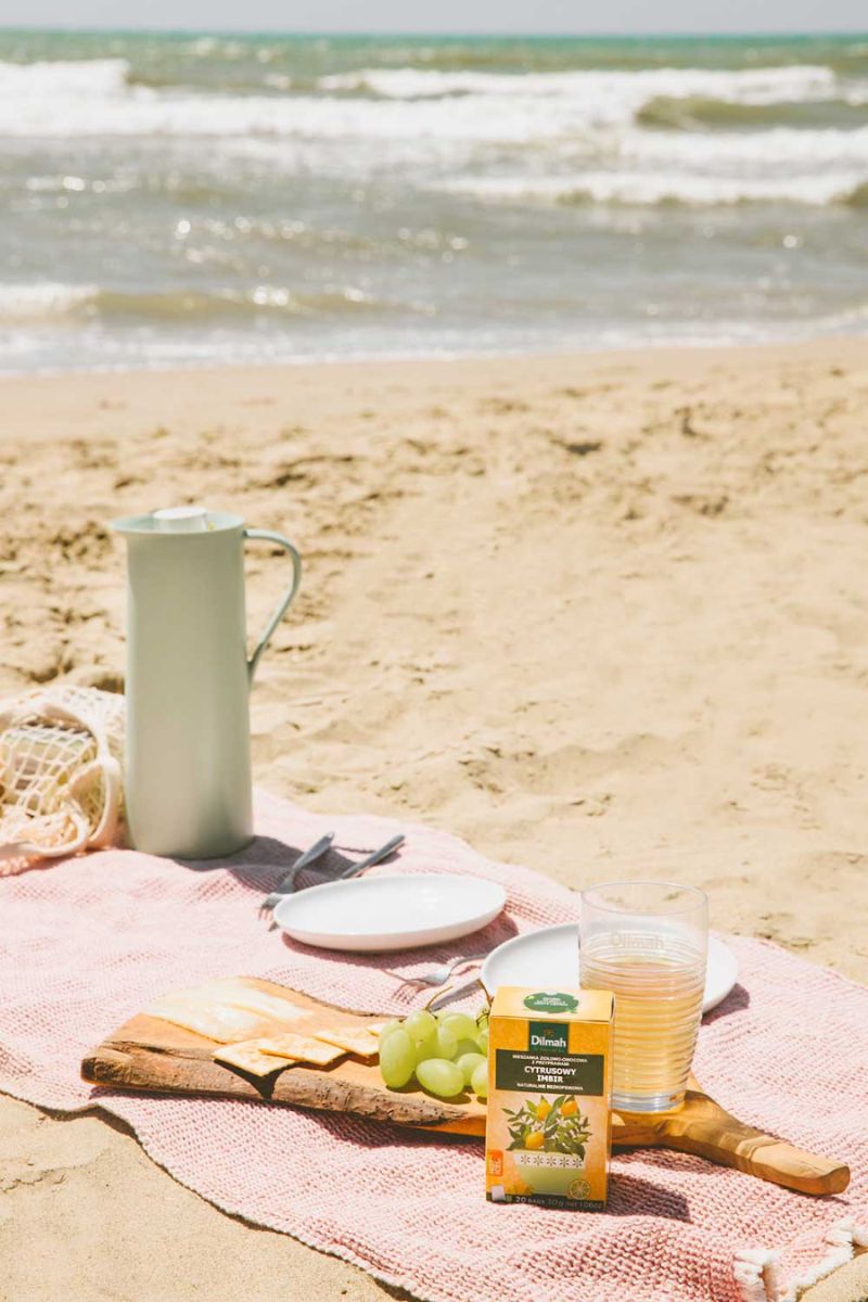 Scena pikniku na plaży. Na kocu, na piasku znajdują się: szklanka z zimnymnapojem, napar cytrynowy Dilmah Naturally Zesty Lemon, deska z serem, krakersami i winogronem oraz sztućce i talerzyki.
