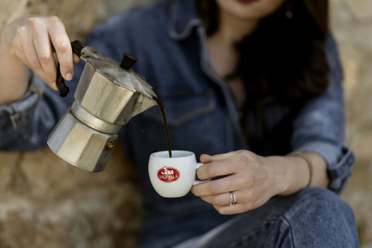 Kobieta nalewająca świeżo zaparzoną w kawiarce kawę Saquella do filiżanki do espresso z logo Saquella.