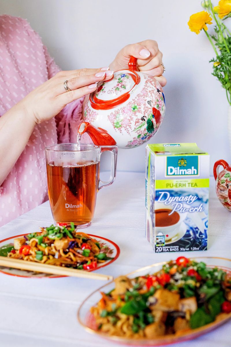 Kobieta nalewająca czerwoną herbatę pu-erh do kubka. Obok orientalnego imbryka pudełko z herbatą Dilmah Dynasty Pu-Erh Tea oraz dania kuchni chińskiej.
