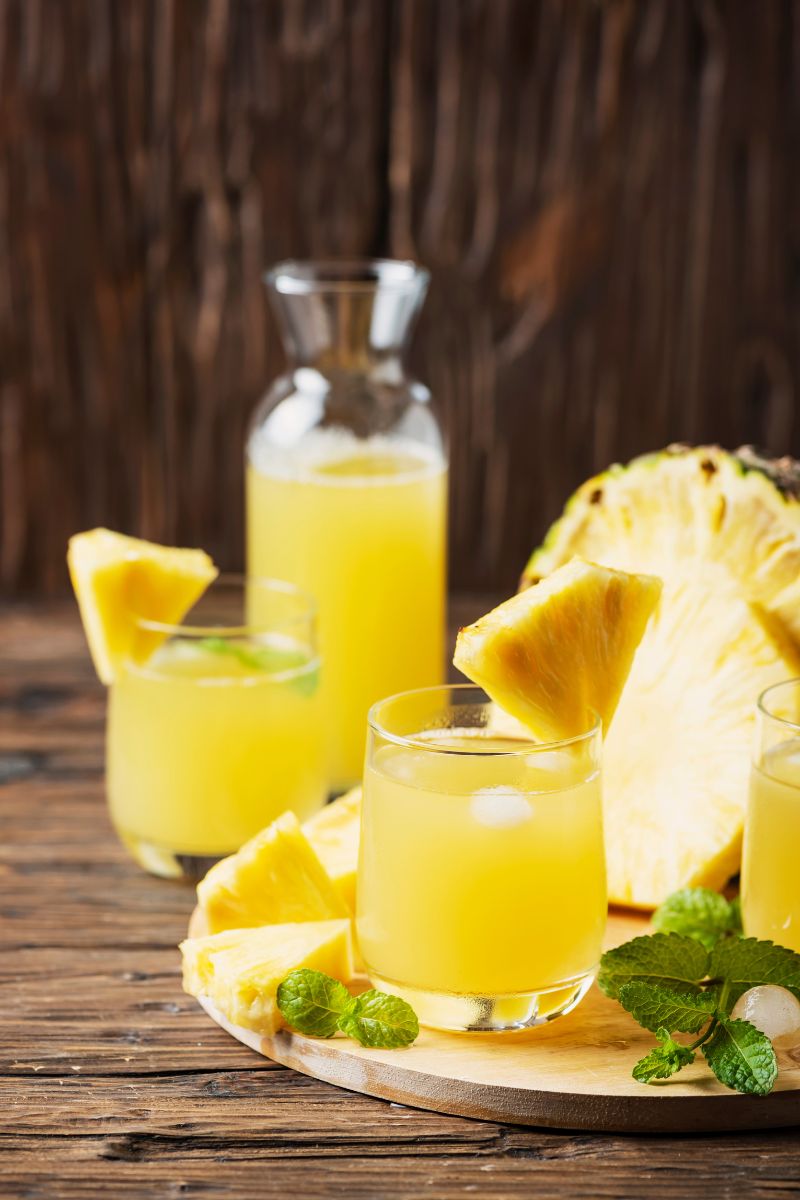 Lemoniada ananasowa w szklance i karafce, stojąca na dębowym stole wśród kawałków ananasa, listków mięty i kostek lodu.