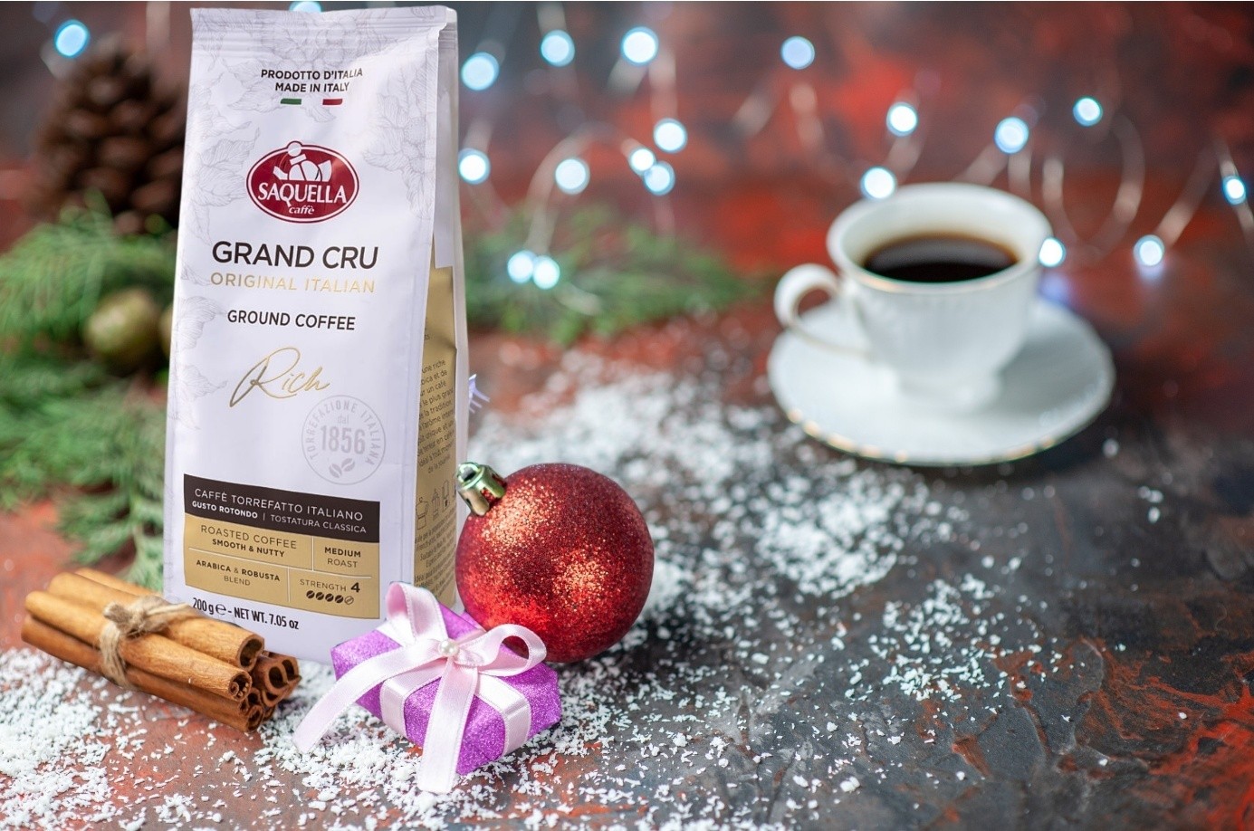 Opakowanie kawy Saquella Grand Cru Rich Original Italian wśród świątecznych ozdób. W tle filiżanka z espresso.
