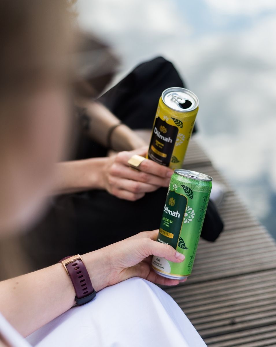 Zbliżenie na puszki herbaty Dilmah Lemon Black Ice Tea i Dilmah Jasmine Flavor Green Ice Tea. Dwie osoby siedzą w plenerze na molo, delektując się orzeźwiającymi napojami. W tle widać spokojną wodę. Letnia atmosfera, która podkreśla relaks i przyjemność wspólnego spędzania czasu na świeżym powietrzu.