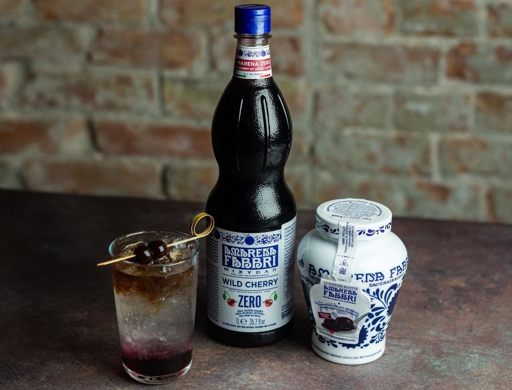 Szklanka Cherry Tonic Espresso Zero z dekoracją z wiśni Amarena Fabbri i kostkami lodu. Obok słoiczek Amarena Fabbri i butelka syropu wiśniowego bez cukru Fabbri Mixybar Amarena Zero.