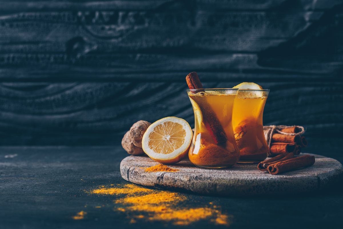 Lemoniada z kardamonem, imbirem i kurkumą w dwóch przezroczystych, tureckichszklankach. Napoje przyozdobione laską cynamonu oraz plastrami cytryny. Obokszklanek znajdują się korzenne przyprawy.
