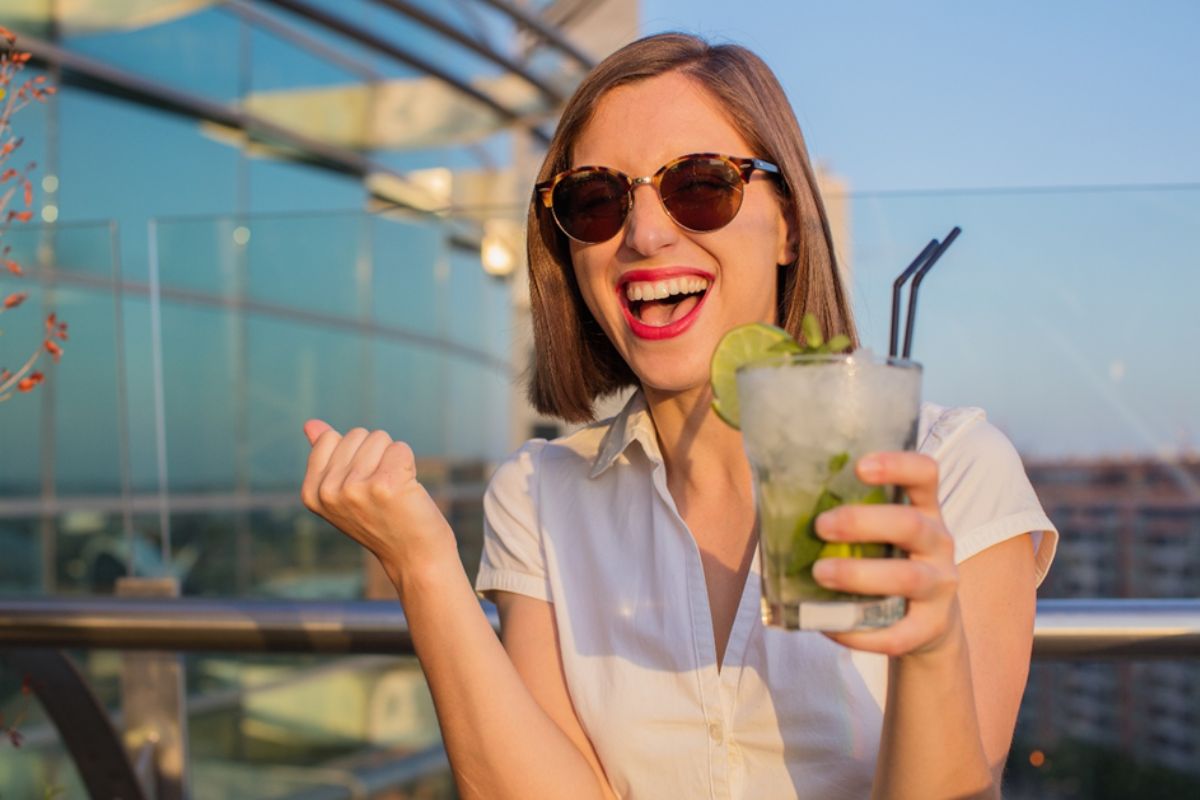 Kobieta w okularach przeciwsłonecznych z dużym uśmiechem na twarzy, która na balkonie popija ulubione bezalkoholowe mojito.