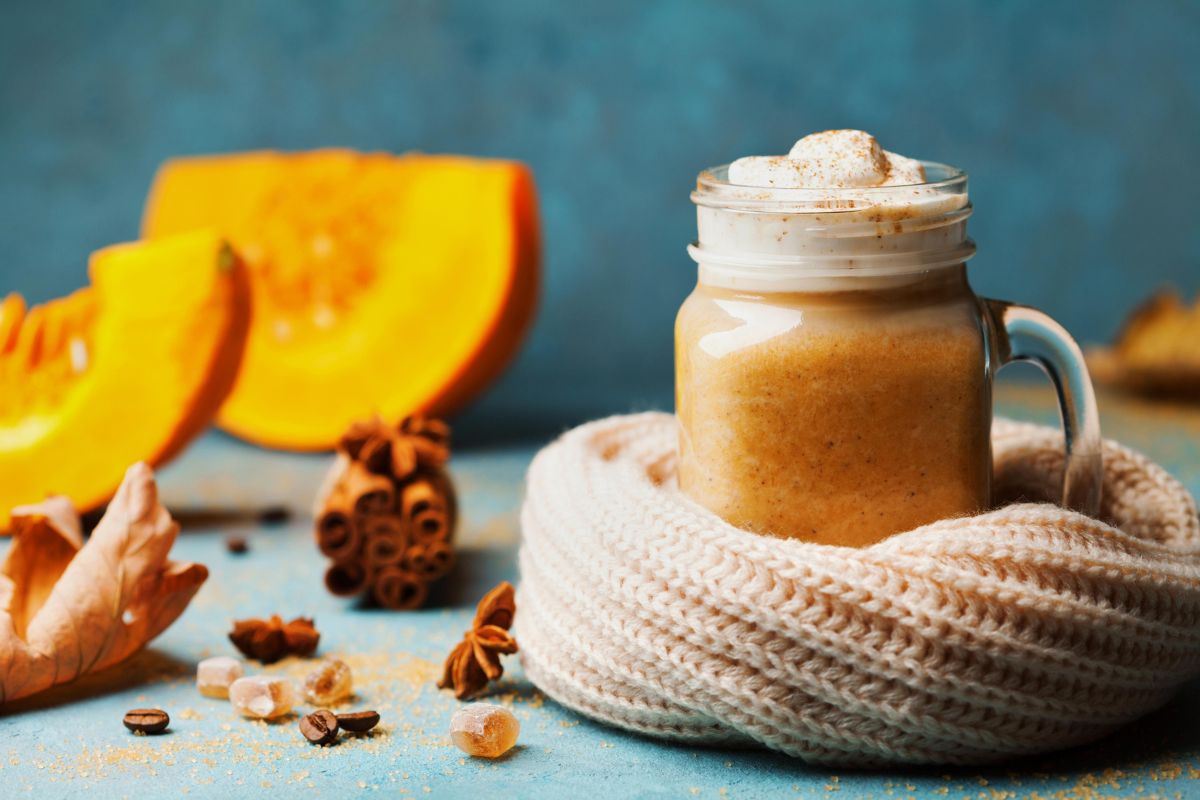 Apetyczne pumpkin spicy latte z piankami marshmallow i cynamonem w kubku otoczonym sweterkiem. W tle dynia, suche jesienne liście i przyprawy korzenne.