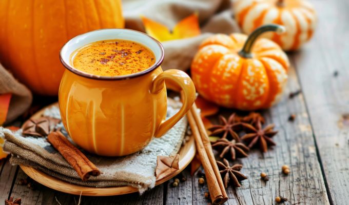 Dyniowa kawa Pumpkin Spice Latte podana w kubku w kształcie dyni. W tle małe dynie dekoracyjne, goździki, laski cynamonu i gwiazdki anyżu.