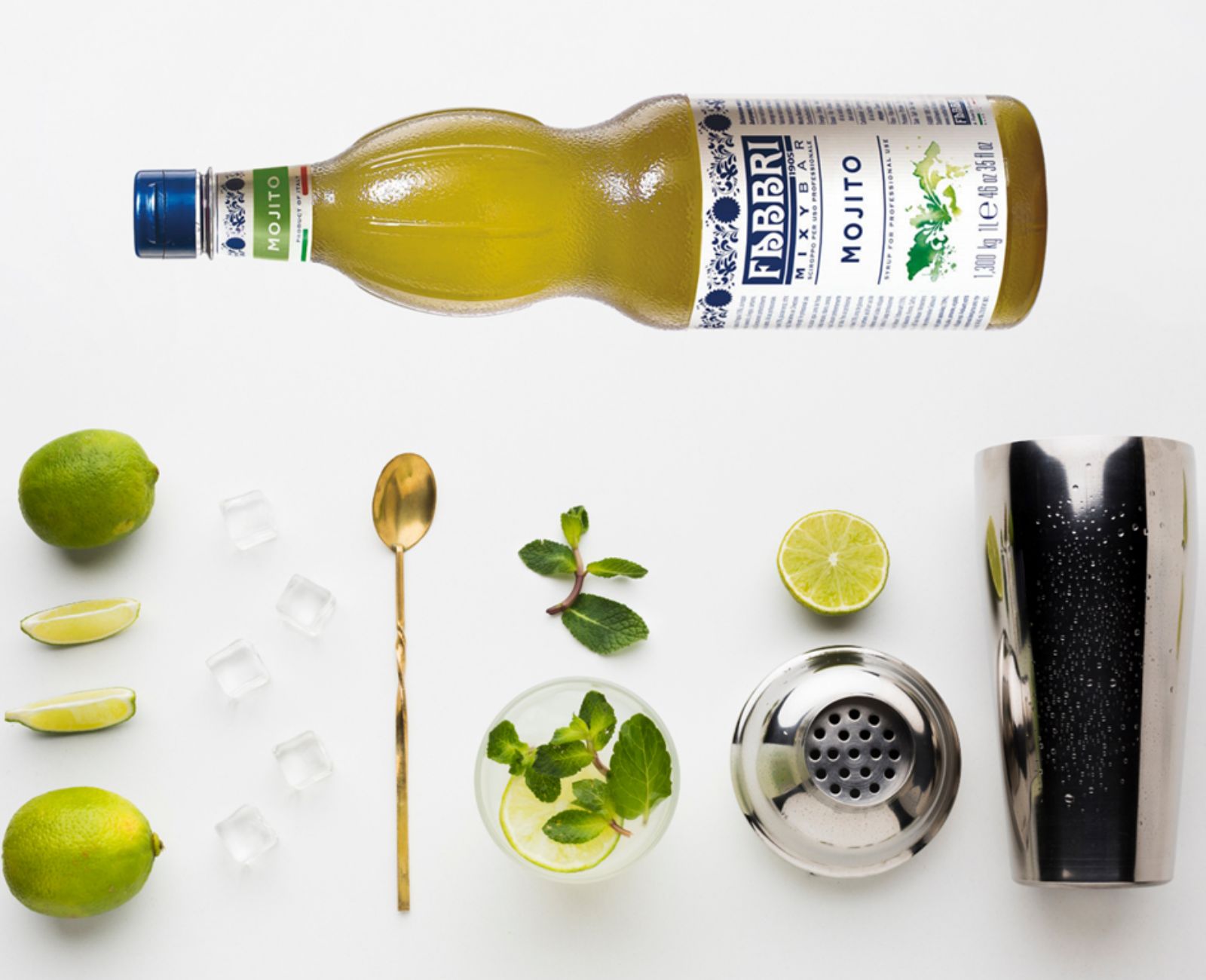 Składniki na Virgin Mojito ułożone na białym stole: świeże limonki, lód, zielone listki mięty, butelka syropu Fabbri Mojito. Obok widoczne są akcesoria barmańskie – szklanka, shaker i długa łyżka do mieszania drinków.
