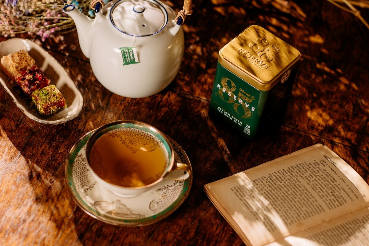 Zielona herbata w torebkach, w eleganckiej puszce Dilmah Ceylon Green Tea with Mint na blacie obok książki, imbryka i filiżanki.