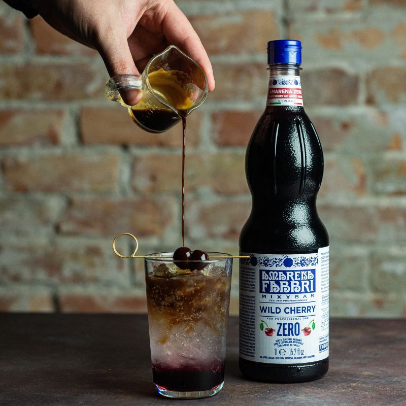 Wlewanie kawy do szklanki z Cherry Tonic Espresso Zero z wiśniami Amarena Fabbri i lodem. Obok butelka syropu wiśniowego bez cukru Fabbri Mixybar Amarena Zero.